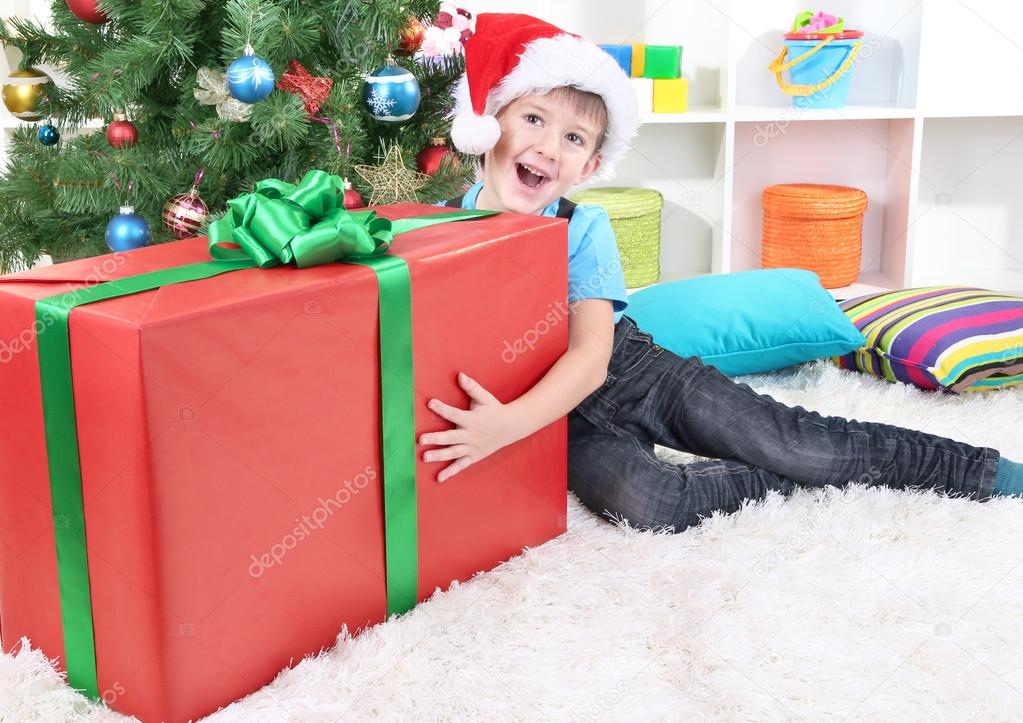Happy boy in Santa hat sits near Christmas tree with big gift in hands