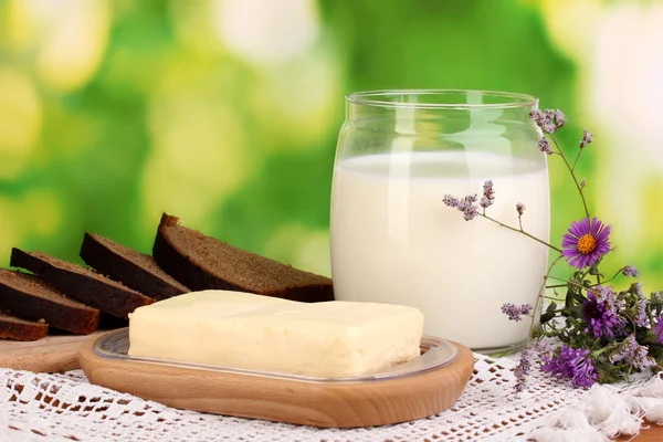 Mantequilla sobre soporte de madera rodeada de pan y leche sobre fondo natural — Foto de Stock