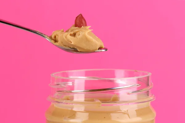 Köstliche Erdnussbutter in Glas und Löffel auf rosa Hintergrund — Stockfoto