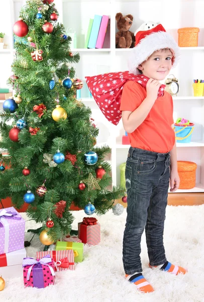 Little boy in Santa hat stands near Christmas tree holding bag with gifts Stock Image