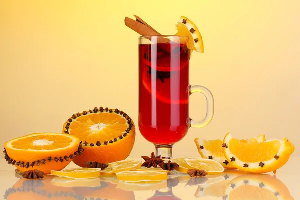 Pachnące grzane wino w szkle z przyprawami i pomarańcze wokół na żółtym tle — Zdjęcie stockowe