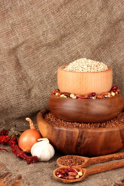 Trigo cru, trigo sarraceno e feijão em tigelas de madeira na mesa sobre fundo de pano de saco — Fotografia de Stock