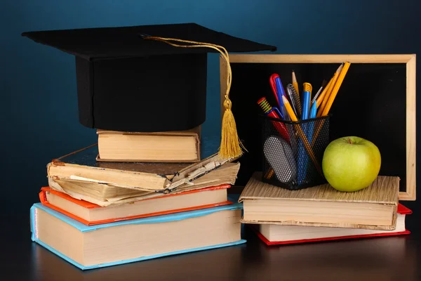 Livros e magister cap contra placa da escola em mesa de madeira no fundo azul — Fotografia de Stock