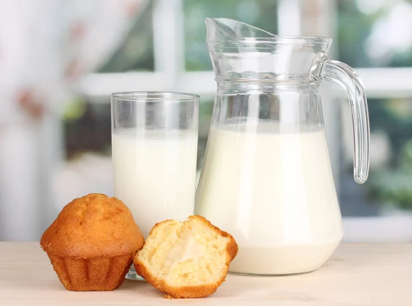 Jarro e copo de leite com muffins na mesa de madeira no fundo da janela — Fotografia de Stock