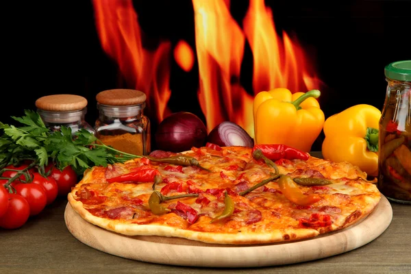 Tasty pepperoni pizza with vegetables on wooden board on flame background — Stock Photo, Image