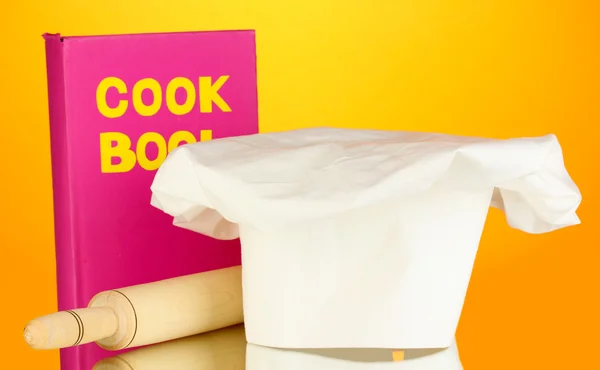 Kochmütze mit Schlachtplatte und Kochbuch auf orangefarbenem Hintergrund — Stockfoto