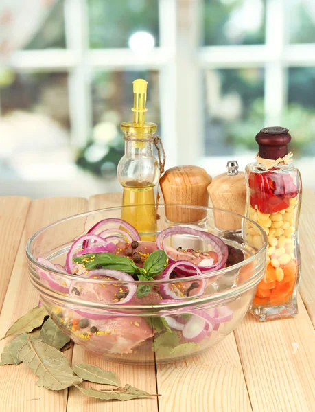 Un gros morceau de porc mariné dans un bol avec des herbes, des épices et de l'huile de cuisson sur une table en bois sur fond de fenêtre — Photo