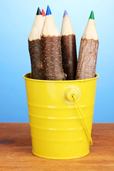 Barevné dřevěné tužky v žlutém kbelíku na dřevěný stůl na modrém pozadí — Stock fotografie