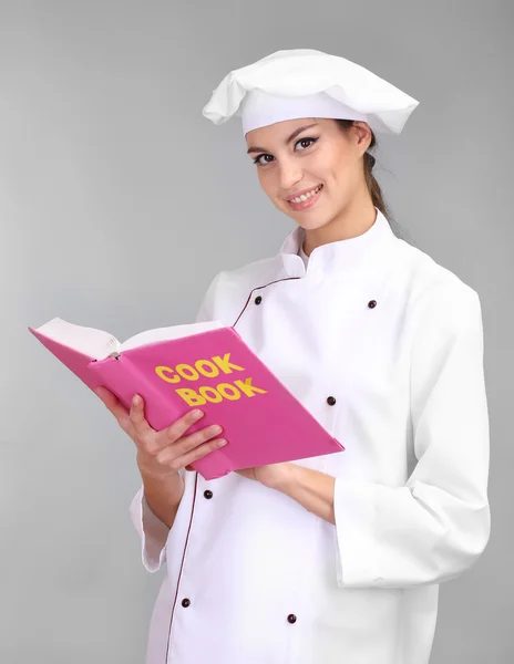 Potret wanita muda koki dengan buku masak di latar belakang abu-abu — Stok Foto