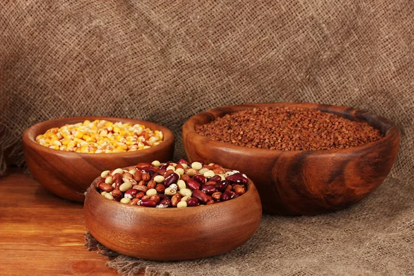 Raw corn,buckwheat and beans in wooden bowls on table on sackcloth background — Stock Photo, Image