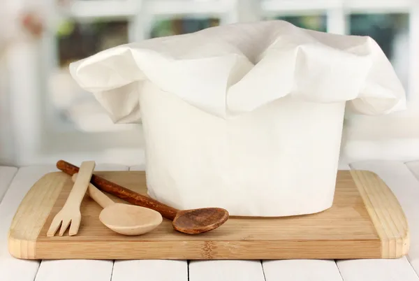 Chef's hat with spoons on board on wooden table on window background — Stock Photo, Image