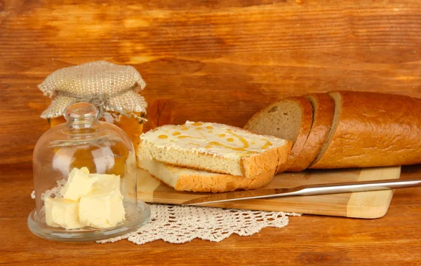 Βούτυρο σε γυάλινο πιατάκι με γυάλινο κάλυμμα και φρέσκο ψωμί, μέλι σε ξύλινα φόντο — Φωτογραφία Αρχείου