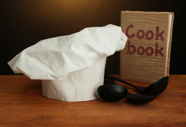 Kucharski kapelusik z łyżki i książka kucharska na stole na brązowym tle — Zdjęcie stockowe