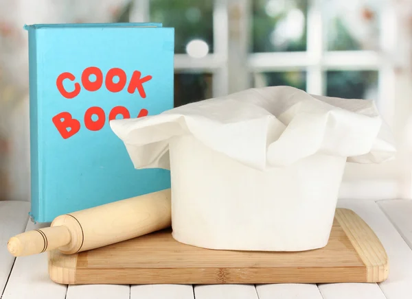 Chef's hat with battledore on board on wooden table on window background — Stock Photo, Image