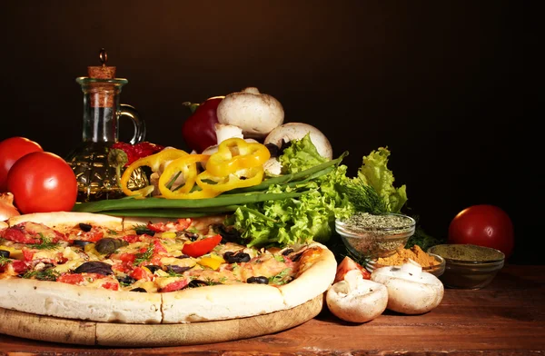 Delicious pizza, vegetables and spices on wooden table on brown background — Stock Photo, Image