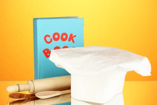 Chef's hat with battledore and cook book on orange background — Stock Photo, Image