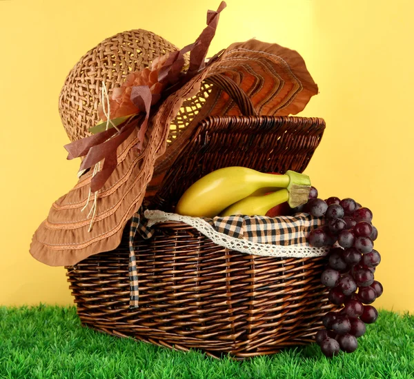 Picnic basket with fruits on grass on yellow background — Stock Photo, Image