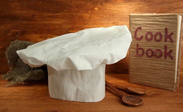 Sombrero de chef con cucharas y libro de cocina sobre fondo de madera — Foto de Stock