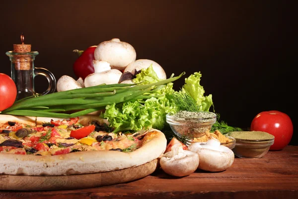 Delicious pizza, vegetables and spices on wooden table on brown background — Stock Photo, Image