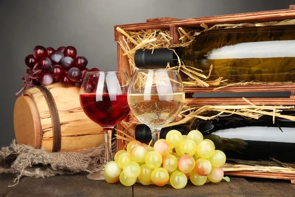 Caixa de madeira com garrafas de vinho, barril, vinho e uva na mesa de madeira em fundo cinza — Fotografia de Stock