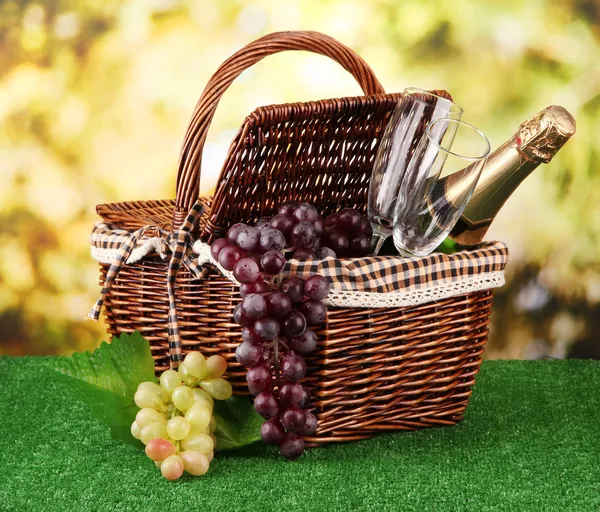 Picnic basket and bottle of champagne on grass on bright background — Zdjęcie stockowe