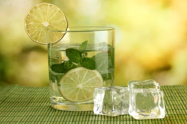 Bicchiere d'acqua con ghiaccio, menta e calce sul tavolo su sfondo luminoso — Foto Stock