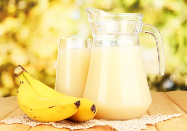 Copo cheio e jarro de suco de banana e bananas na mesa de madeira ao ar livre — Fotografia de Stock