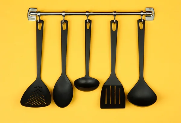 Black kitchen utensils on silver hooks, on yellow background — Stock Photo, Image