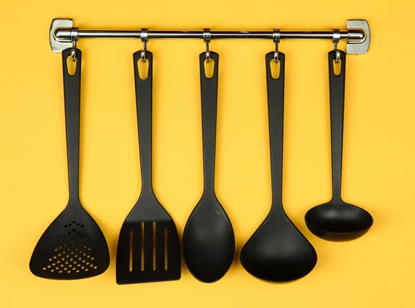 Black kitchen utensils on silver hooks, on yellow background — Stock Photo, Image