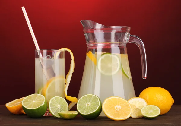 Citrusovou limonádu v džbán a sklenice citrusových kolem na dřevěný stůl na červeném pozadí — Stock fotografie