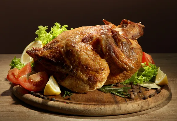 Whole roasted chicken with vegetables, on wooden table, on brown background — Stock Photo, Image