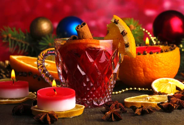 Fragrant mulled wine in glass with spices and oranges around on wooden table on red background — Stock Photo, Image