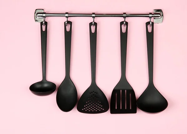 Black kitchen utensils on silver hooks, on pink background — Stock Photo, Image