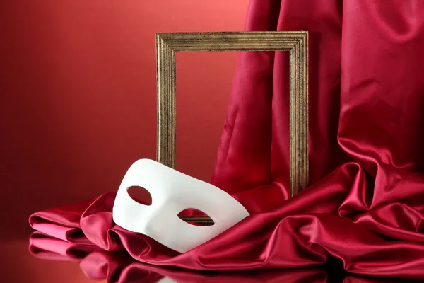 White mask, empty frame and golden silk fabric, on red background — Stock Photo, Image