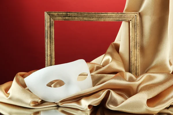 White mask, empty frame and golden silk fabric, on red background — Stock Photo, Image