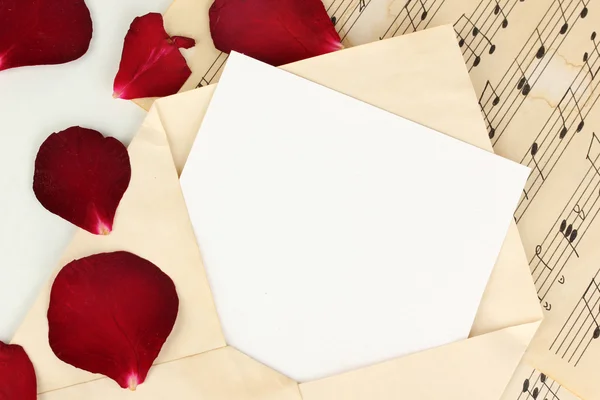 Old envelope with blank paper and dried rose petals on music sheets close up — Stock Photo, Image