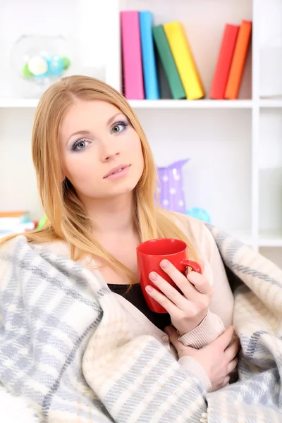 Attraktive junge Frau sitzt auf dem Sofa, hält Tasse mit Heißgetränk, auf dem heimischen Innenhintergrund — Stockfoto