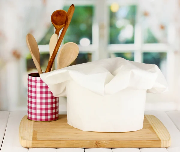 Šéfkuchařské čepice s lžíce na palubě na dřevěný stůl na pozadí okna — Stock fotografie