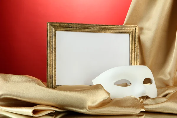 Máscara branca, moldura vazia e tecido de seda dourada, sobre fundo vermelho — Fotografia de Stock