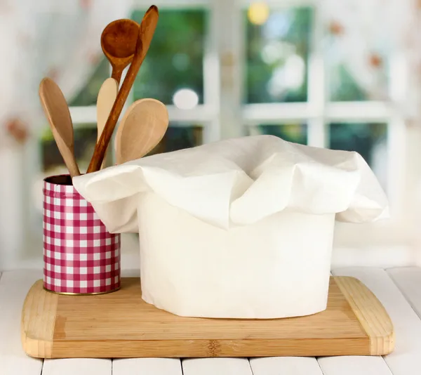 Chef's hat with spoons on board on wooden table on window background — Stock Photo, Image