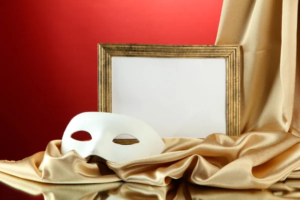 White mask, empty frame and golden silk fabric, on red background — Stock Photo, Image