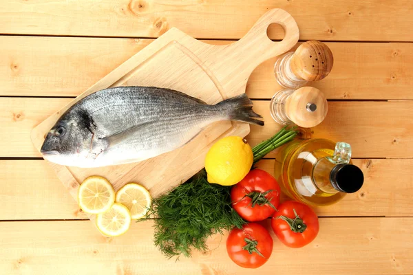 Fresh dorado on chopping board with lemon and vegetables on wooden table — Stock Photo, Image