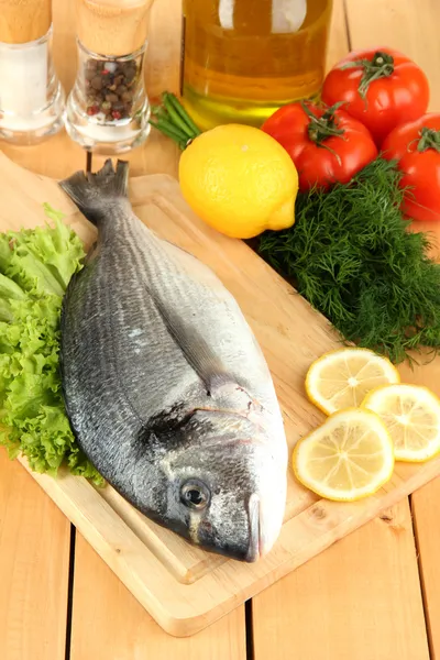 Fresh dorado on chopping board with lemon and vegetables on wooden table — Stock Photo, Image