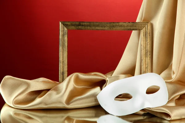 White mask, empty frame and golden silk fabric, on red background — Stock Photo, Image