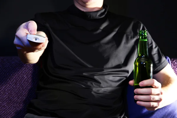Main d'homme tenant une télécommande TV et une bouteille de bière, sur fond sombre — Photo