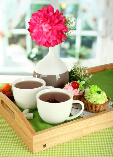 Cups of tea with flower and cakes on wooden tray on table in room — Stock Photo, Image