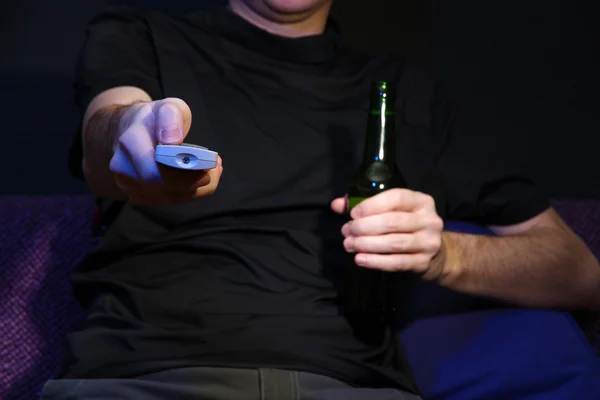 Mano de hombre sosteniendo un mando a distancia TV y botella de cerveza, sobre fondo oscuro — Foto de Stock