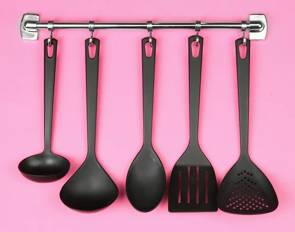 Black kitchen utensils on silver hooks, on pink background — Stock Photo, Image