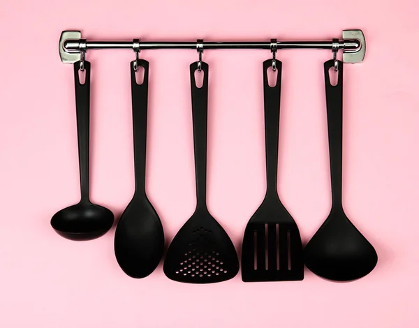Black kitchen utensils on silver hooks, on pink background — Stock Photo, Image