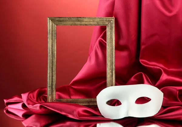 Máscara branca, moldura vazia e tecido de seda dourada, sobre fundo vermelho — Fotografia de Stock
